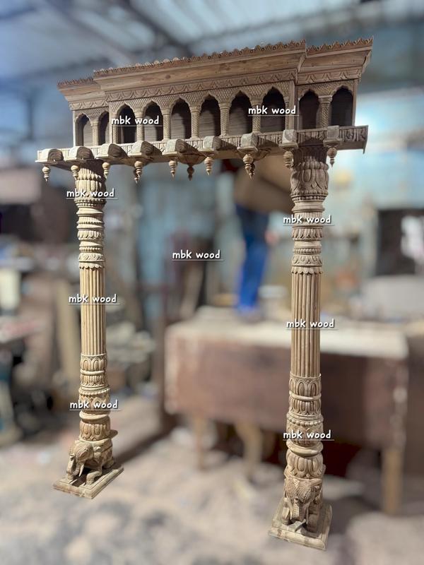 Classic Pooja Mandir Side Design Description:This design features two elaborately carved Chettinad-style wooden pillars supporting a grand top canopy. The pillars have vertical fluted patterns with layered ornamental rings at intervals, leading down to detailed elephant carvings at the base, symbolizing strength and auspiciousness. The top canopy has a series of arched niches with miniature columns, giving a temple gopuram-inspired appearance. Beneath the arches, lotus bud hanging motifs add traditional grace. The roof edge has a scalloped pattern, adding depth and elegance to the structure. The overall design exudes royal South Indian temple aesthetics, making it perfect for a heritage-style pooja mandir or a traditional interior focal point.
